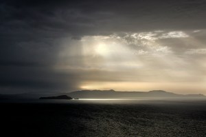 A serene seascape with dark clouds looming overhead, pierced by rays of light breaking through, illuminating a distant island and reflecting on the water below.