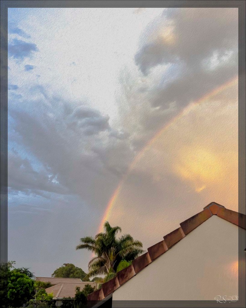 A soft rainbow arches across a cloudy sky at sunset, above a suburban rooftop and palm trees.