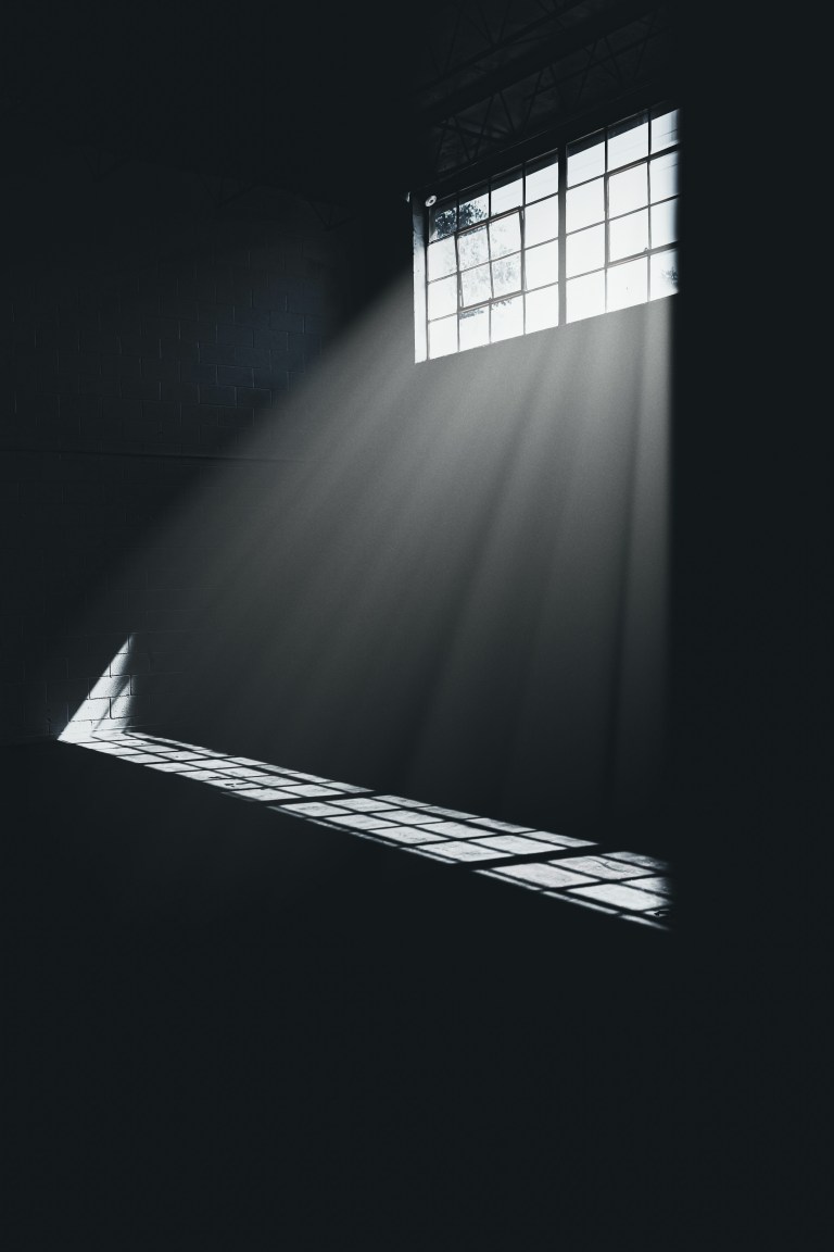Light streaming through a high window into a dark room — symbol of grace entering even in surrender.