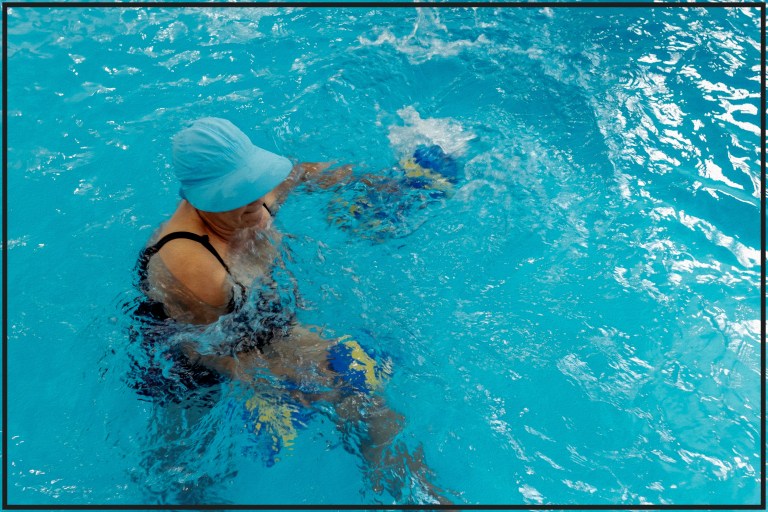 Older woman in blue swim cap doing water aerobics, symbolizing joy in movement and healing before perfection.