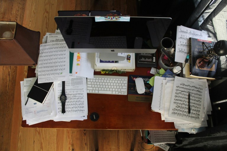 A cluttered wooden desk scattered with sheet music, books, notebooks, and coffee mugs around a computer — a candid glimpse of a restless mind at work.