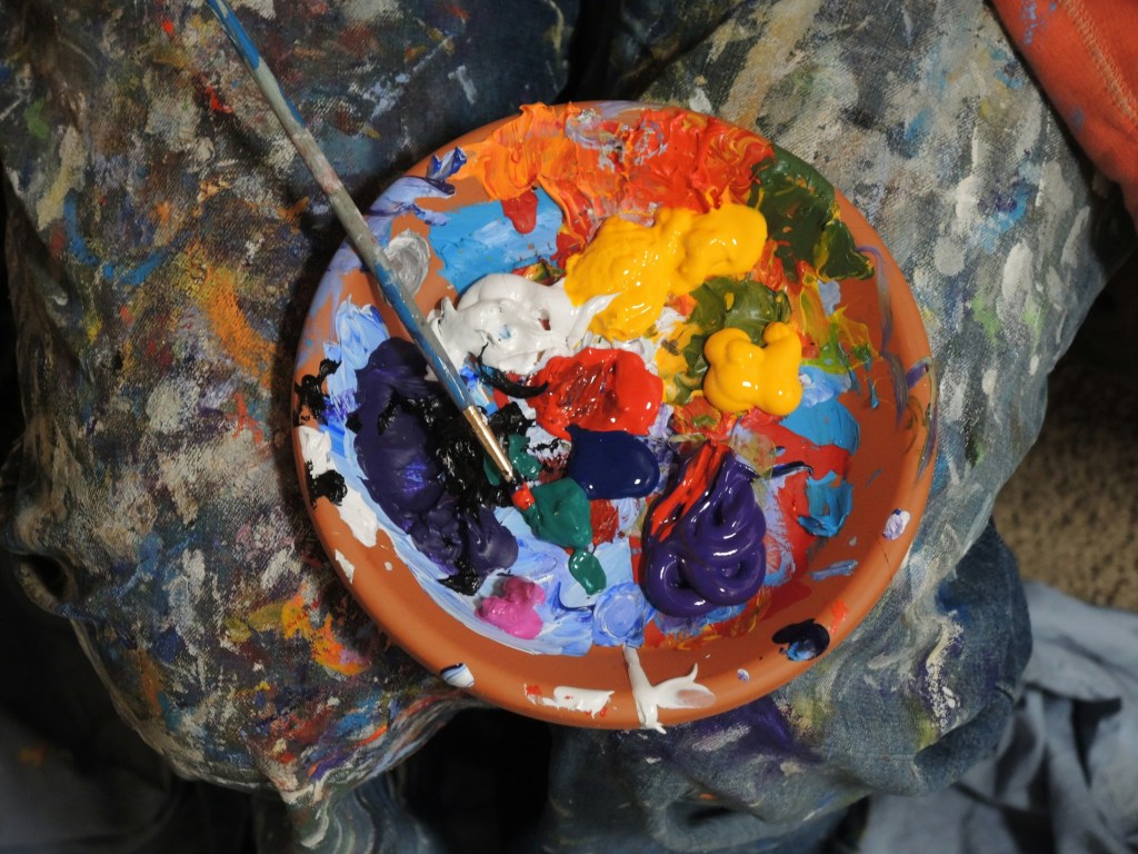 A paint-smeared palette resting on a lap covered in splattered jeans, filled with bold, unblended colors—celebrating the beauty of imperfection.