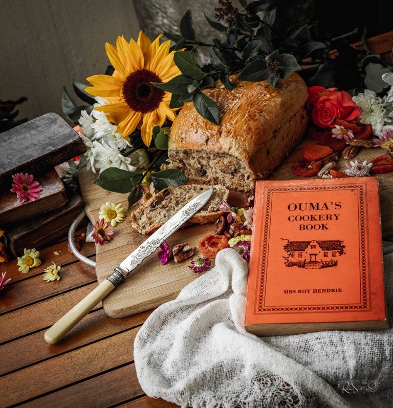 Homemade loaf of seed bread with vintage cookbook, flowers, and rustic knife on a wooden table.