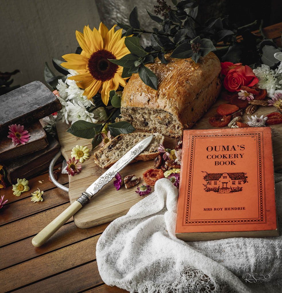 Homemade loaf of seed bread with vintage cookbook, flowers, and rustic knife on a wooden table.