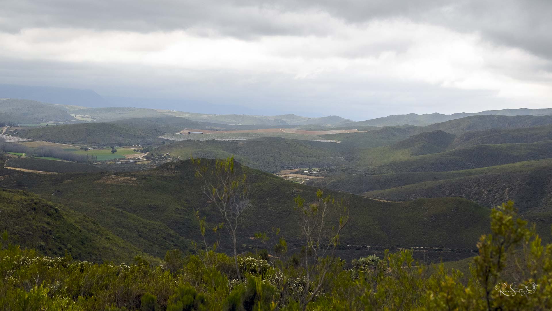 Expansive view of the Klein Karoo with layered green hills, distant farmlands, and dramatic clouds rolling over the horizon.