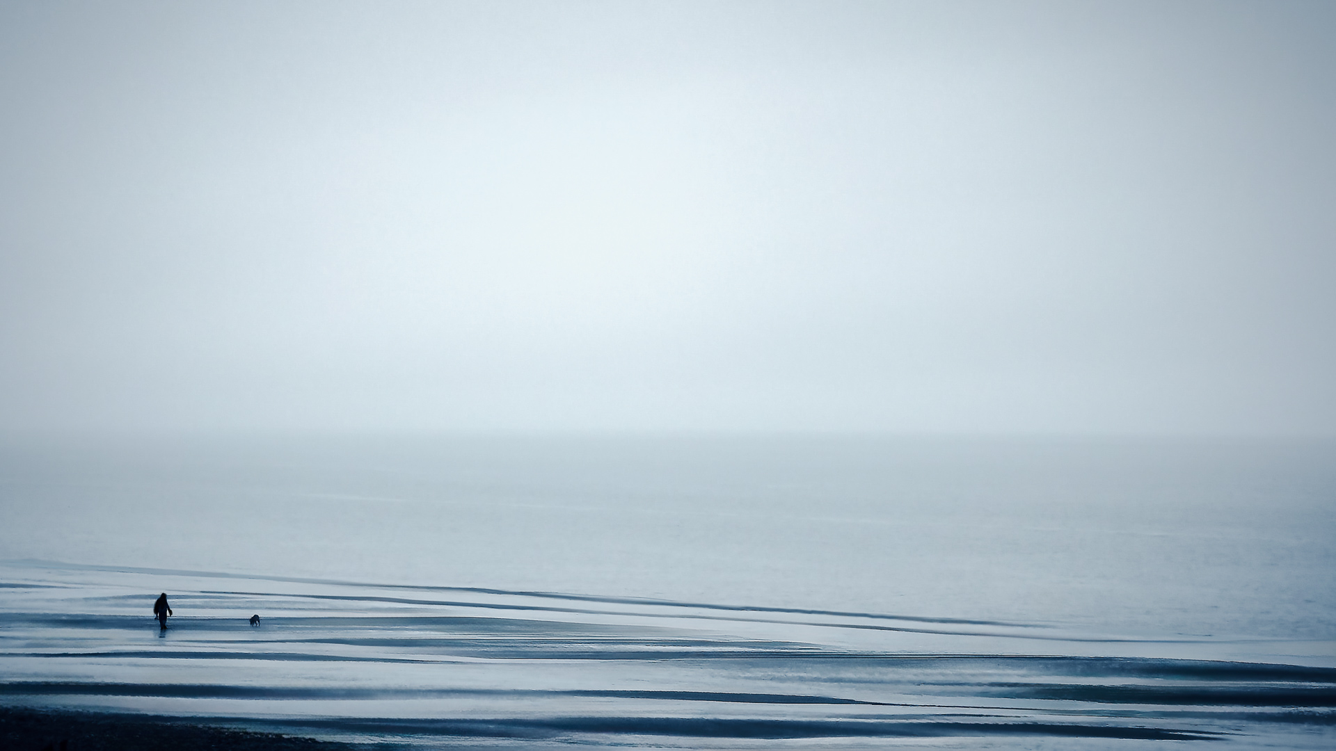A lone figure walks along a frozen shoreline in Alaska beneath a vast, pale sky, surrounded by still water and winter silence.