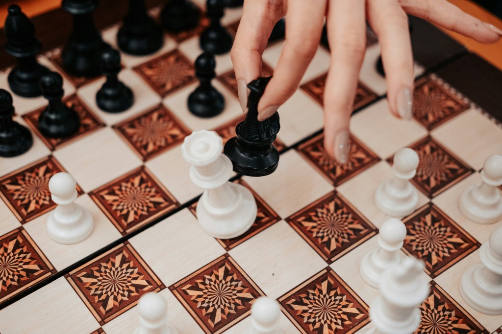 Close-up of a hand moving a black chess piece on a patterned wooden chessboard during a game.
