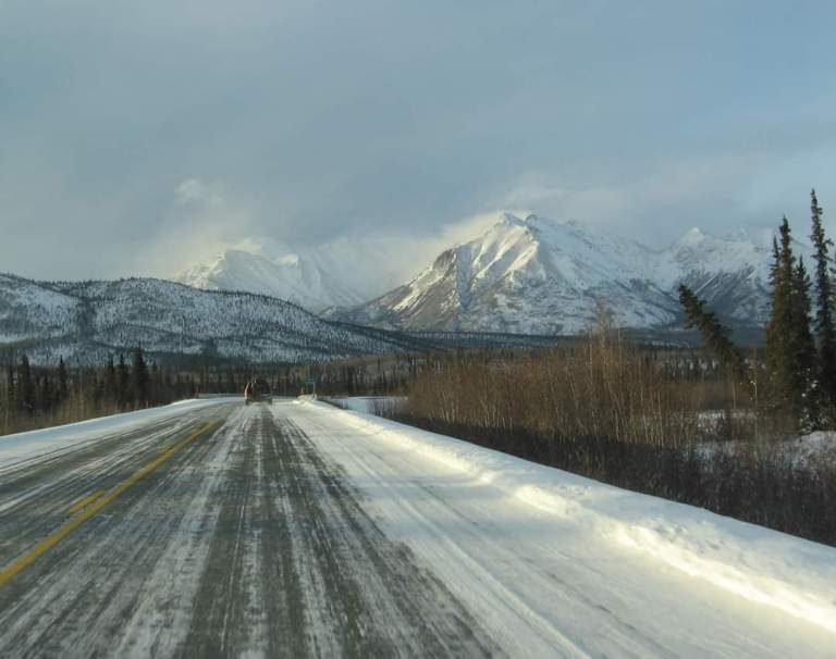 Snow-covered road leading toward sunlit mountains beneath a pale winter sky in Alaska.