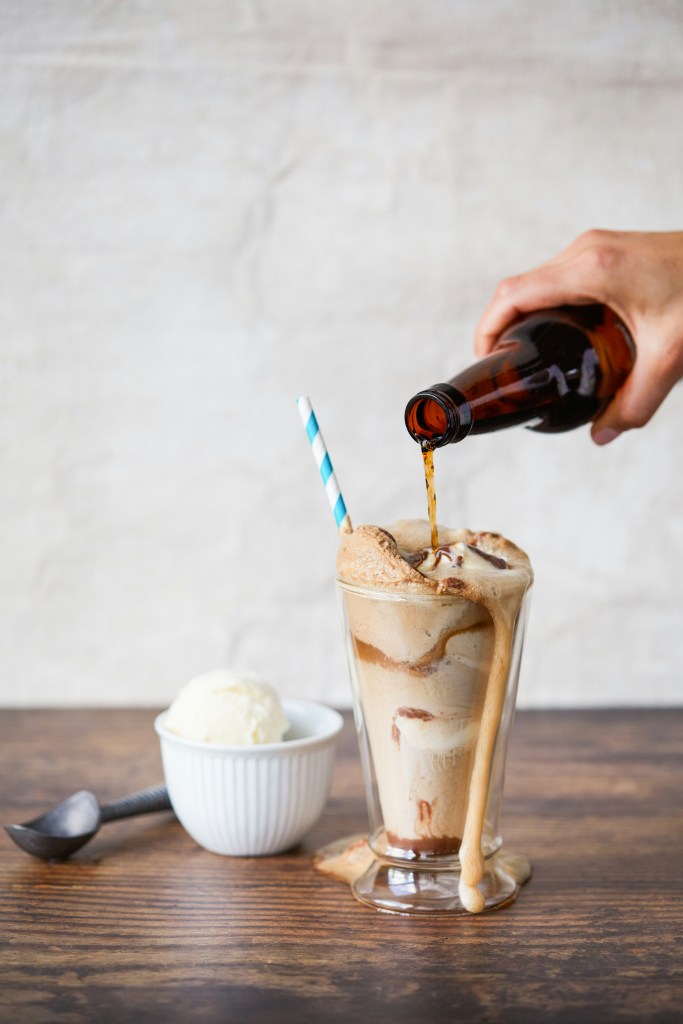 A hand pours root beer from a brown bottle over vanilla ice cream in a tall glass, creating a foamy root beer float.