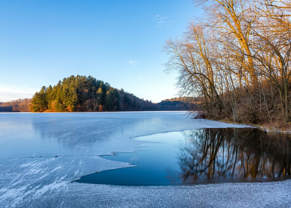 A partially frozen lake reflects bare winter trees beneath a clear blue sky, with golden light touching the shoreline.