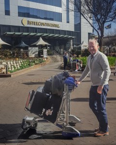 Patrick standing beside a luggage trolley holding three suitcases outside OR Tambo International Airport in Johannesburg.