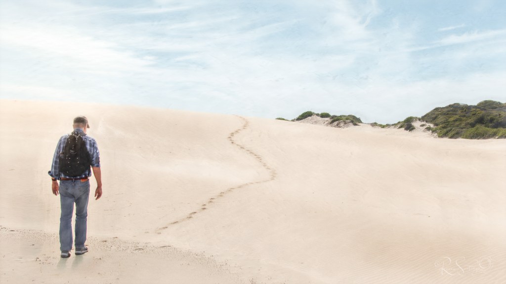 A solitary man walks across pale sand dunes, following a winding trail of footprints beneath a soft blue sky.